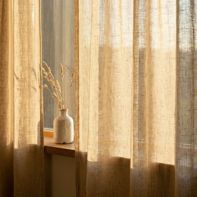 Dettaglio di tende in lino grezzo che filtrano la luce naturale creando un bagliore dorato e soffuso, con piccolo vaso in ceramica sul davanzale
